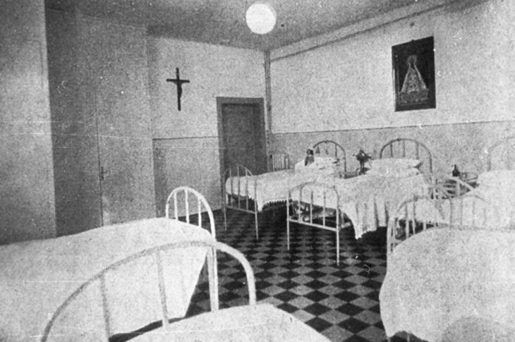 Dormitorio ordinato con letti in ferro, pavimento a scacchi e simboli religiosi